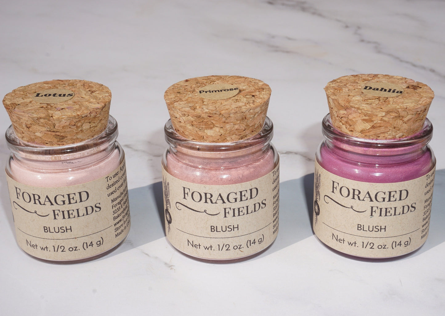 May include: Three glass jars with cork lids containing pink blush powder. The jars are labeled 'Foraged Fields Blush' with the names 'Lotus', 'Primrose', and 'Dahlia' on the lids. The jars are on a white marble surface.