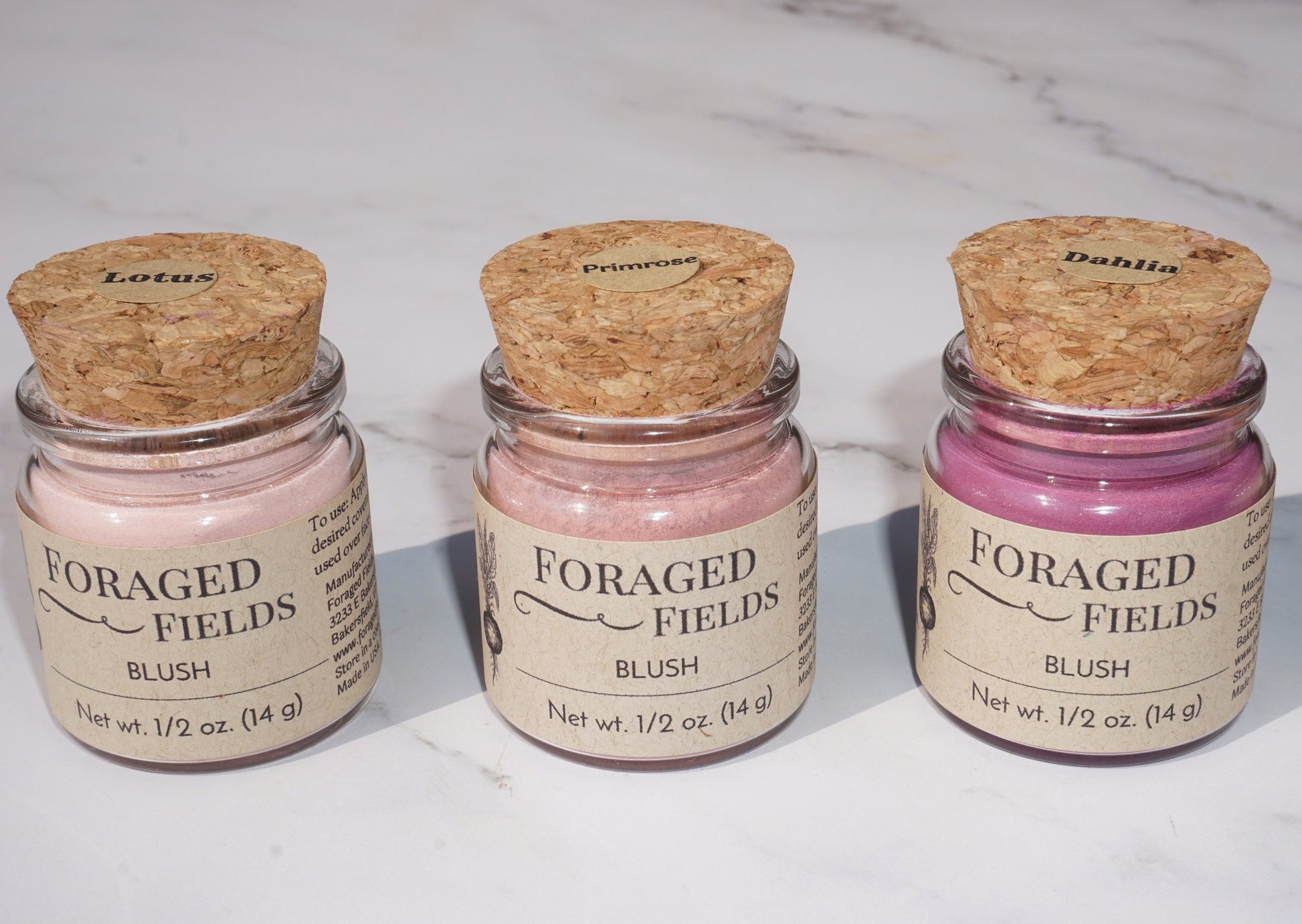 May include: Three glass jars with cork lids containing pink blush powder. The jars are labeled &#39;Foraged Fields Blush&#39; with the names &#39;Lotus&#39;, &#39;Primrose&#39;, and &#39;Dahlia&#39; on the lids. The jars are on a white marble surface.