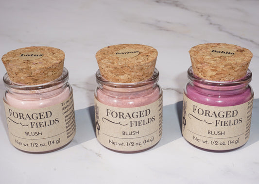 May include: Three glass jars with cork lids containing pink blush powder. The jars are labeled &#39;Foraged Fields Blush&#39; with the names &#39;Lotus&#39;, &#39;Primrose&#39;, and &#39;Dahlia&#39; on the lids. The jars are on a white marble surface.